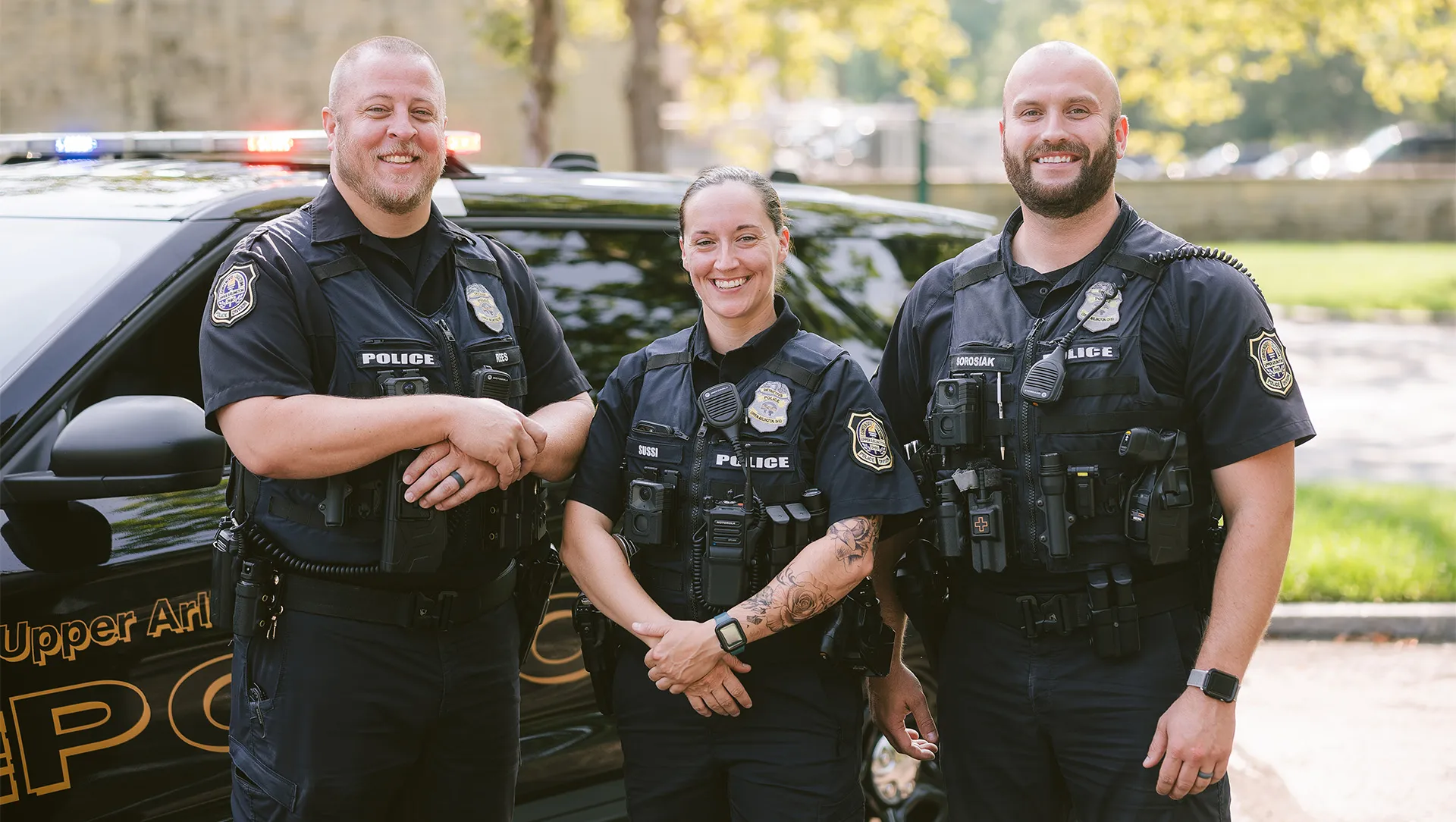Three smiling UAPD police officers in uniform stand in front of a police cruiser.