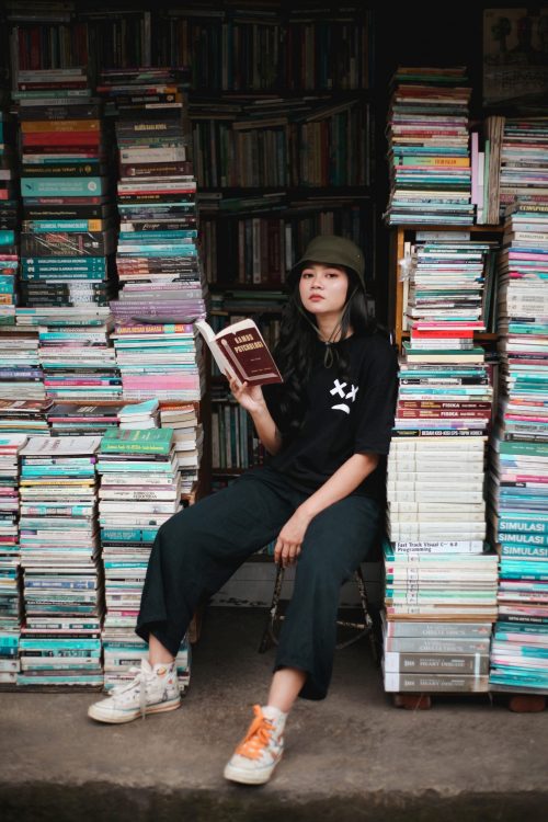 Teen on a stool holding a book, surrounded by tall stacks of books